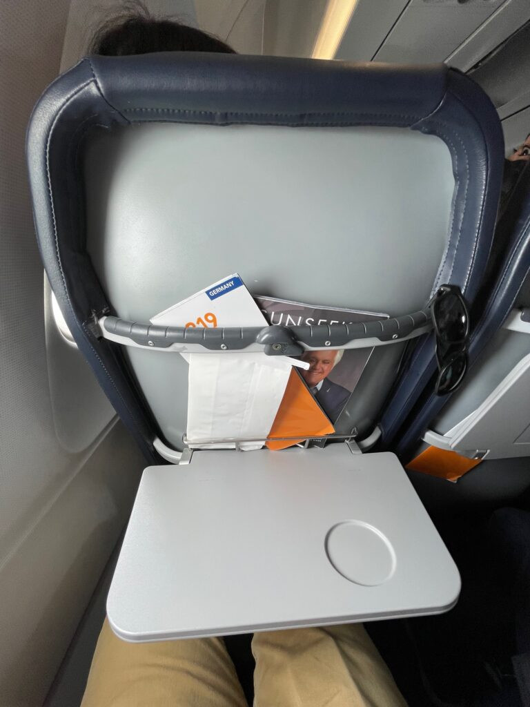 Tray table open on an aircraft seat, showing the literature pocket with a safety card and in-flight magazine.