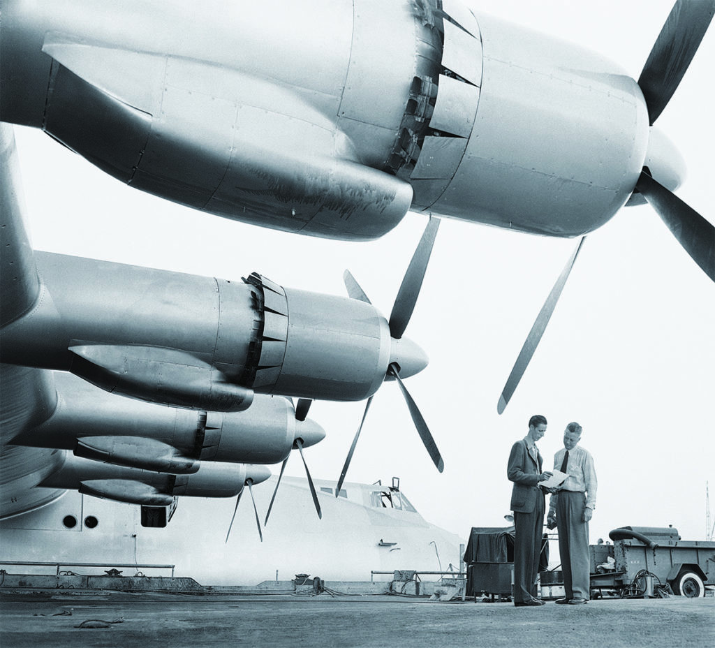 Engineers inspecting the massive Pratt &amp; Whitney engines of the H-4 Hercules, emphasizing the power and complexity of the aircraft.