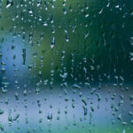 A piece of glass is covered in rain drops with blurred green and blue in the background, representing looking out an airplane window during rain and relating to the question of whether rain cancels flights.