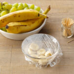 rotten fruit trap method. bowl of rotten banana slices in a bowl covered with plastic wrap that has had toothpick holes poked into it. wood surface background with bananas and grapes nearby.