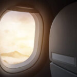 An airplane is visible through an airplane window, with mountains seen in the distance.
