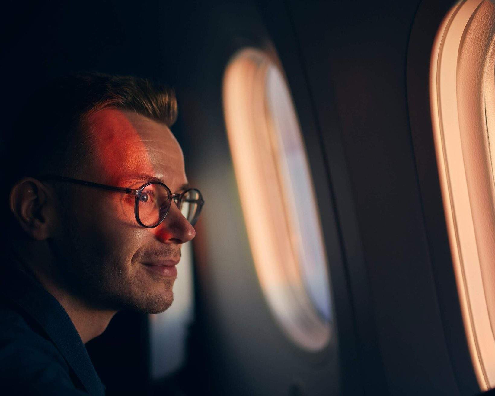 A smiling man wearing glasses looks out of an airplane window, enjoying the view during his domestic flight.