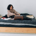 A woman with curly hair is carefully packing clothes into a brown suitcase on a bed, preparing for her trip.