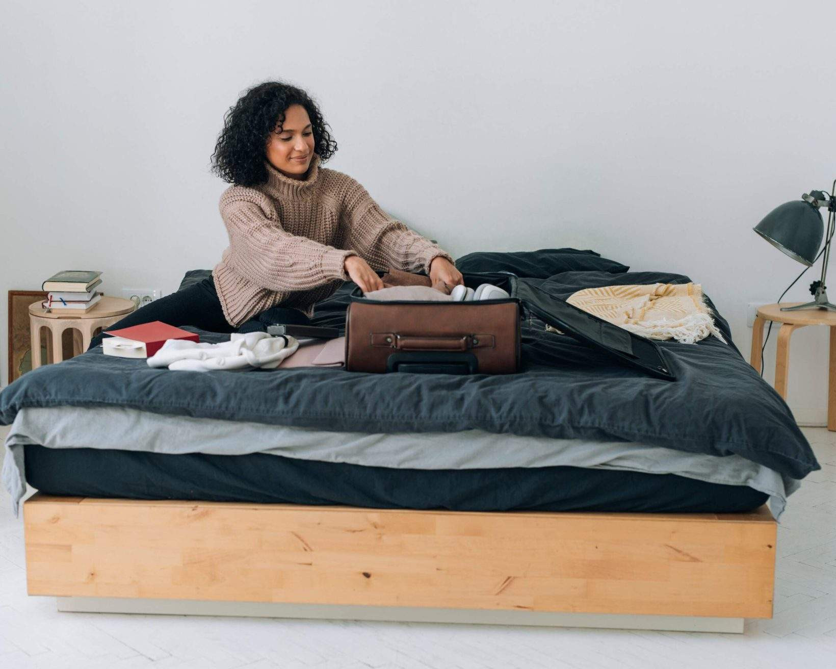 A woman with curly hair is carefully packing clothes into a brown suitcase on a bed, preparing for her trip.