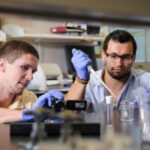 Hardaway, holding video camera, and graduate student Rami Ajjuri work inside a UA lab, conducting research on fruit flies to understand autism spectrum disorders, utilizing model organisms in a science and engineering complex lab environment