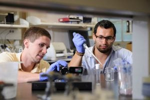 Hardaway, holding video camera, and graduate student Rami Ajjuri work inside a UA lab, conducting research on fruit flies to understand autism spectrum disorders, utilizing model organisms in a science and engineering complex lab environment