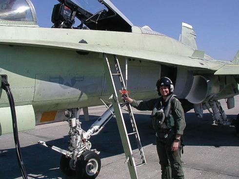 A woman wearing a U.S. Navy flightsuit and helmet stands in front of a ladder leading to the cockpit of a military jet fighter aircraft.