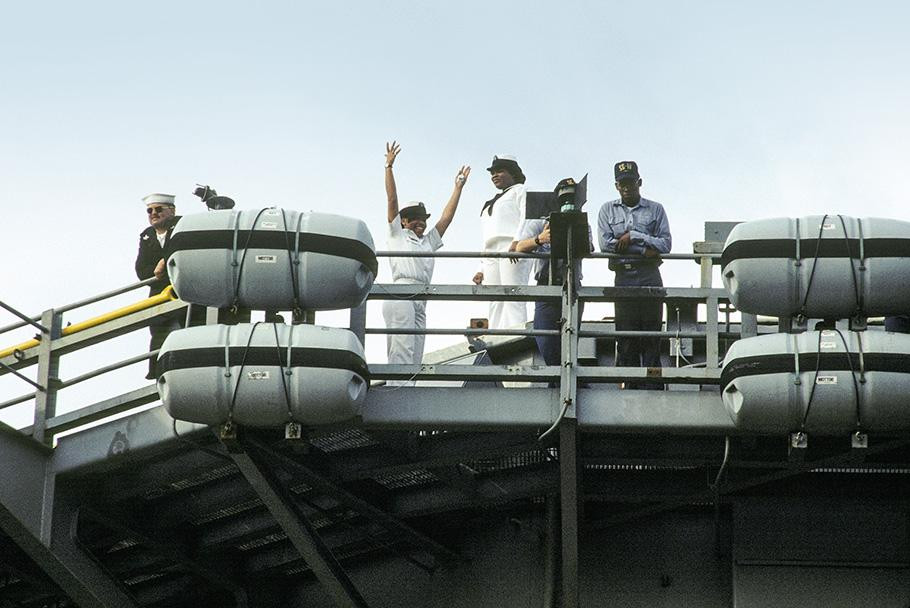 Four U.S. Navy personnel--two women, one man and one person obscured by a map, all in uniform--stand on the deck of an aircraft carrier. The woman on the far left has her hands raised in the air.