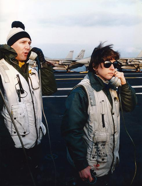 A man and a woman stand on the flight deck of a U.S. Navy aircraft carrier during daylight. Both are wearing white vests and both are speaking on telephones. The wind on the flight deck blows their hair.
