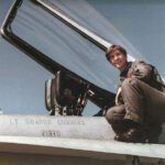 A young woman wearing a U.S. Navy olive-green flightsuit and black combat boots perches next to the cockpit of her F/A-18C miltary jet.
