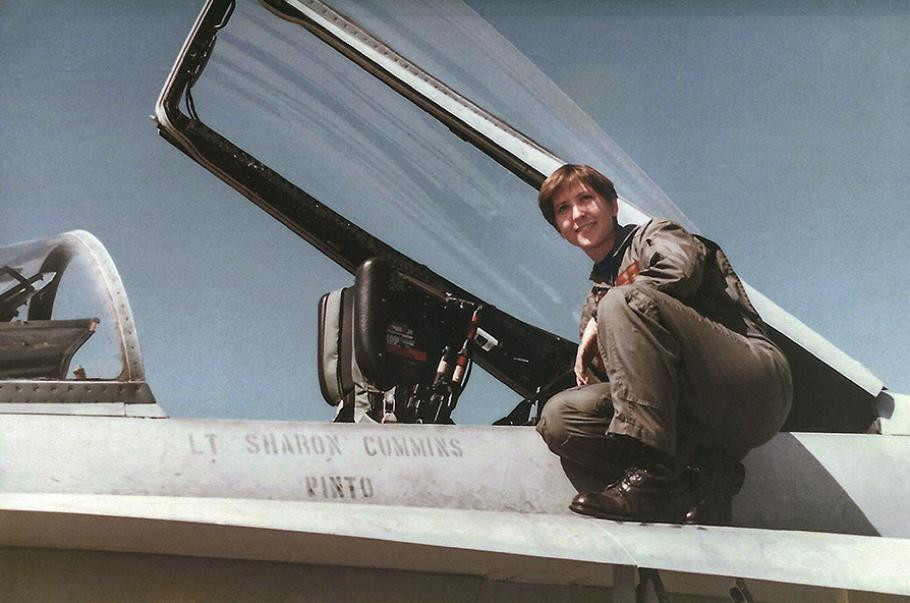 A young woman wearing a U.S. Navy olive-green flightsuit and black combat boots perches next to the cockpit of her F/A-18C miltary jet.