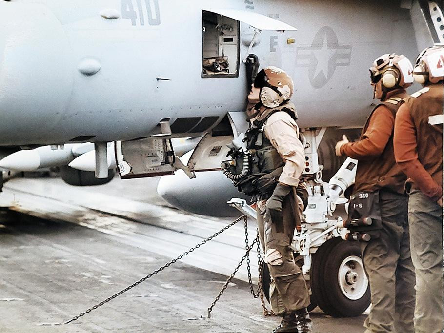 A young woman wearing a U.S. Navy olive-green flightsuit, helmet, goggles, and gloves peers into an open compartment on the side of a gray military jet with U.S. Navy markings.