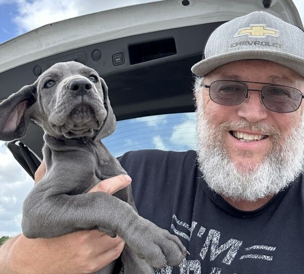 A bearded man named Jason Luck, wearing glasses and a Chevrolet cap, smiles while holding a floppy-eared gray puppy. They are standing outdoors with a car trunk open in the background, hinting at their travel or pet transport plans.