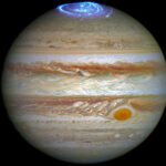 Jupiter against the black backdrop of space. Horizontal bands of orange, brown and beige cover the planet. Blue ribbons of light are auroras at the top of the planet and the Great Red Spot can be seen towards the lower right of the image as a rusty red circle.