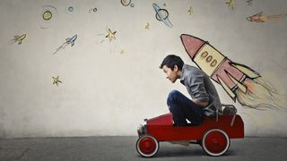 Photograph of a person sitting in a red toy car in front of a gray wall with spaceships and planets drawn on.