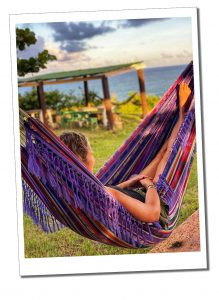 A blonde woman sitting a purple hammock looking out to sea on a sunny evening